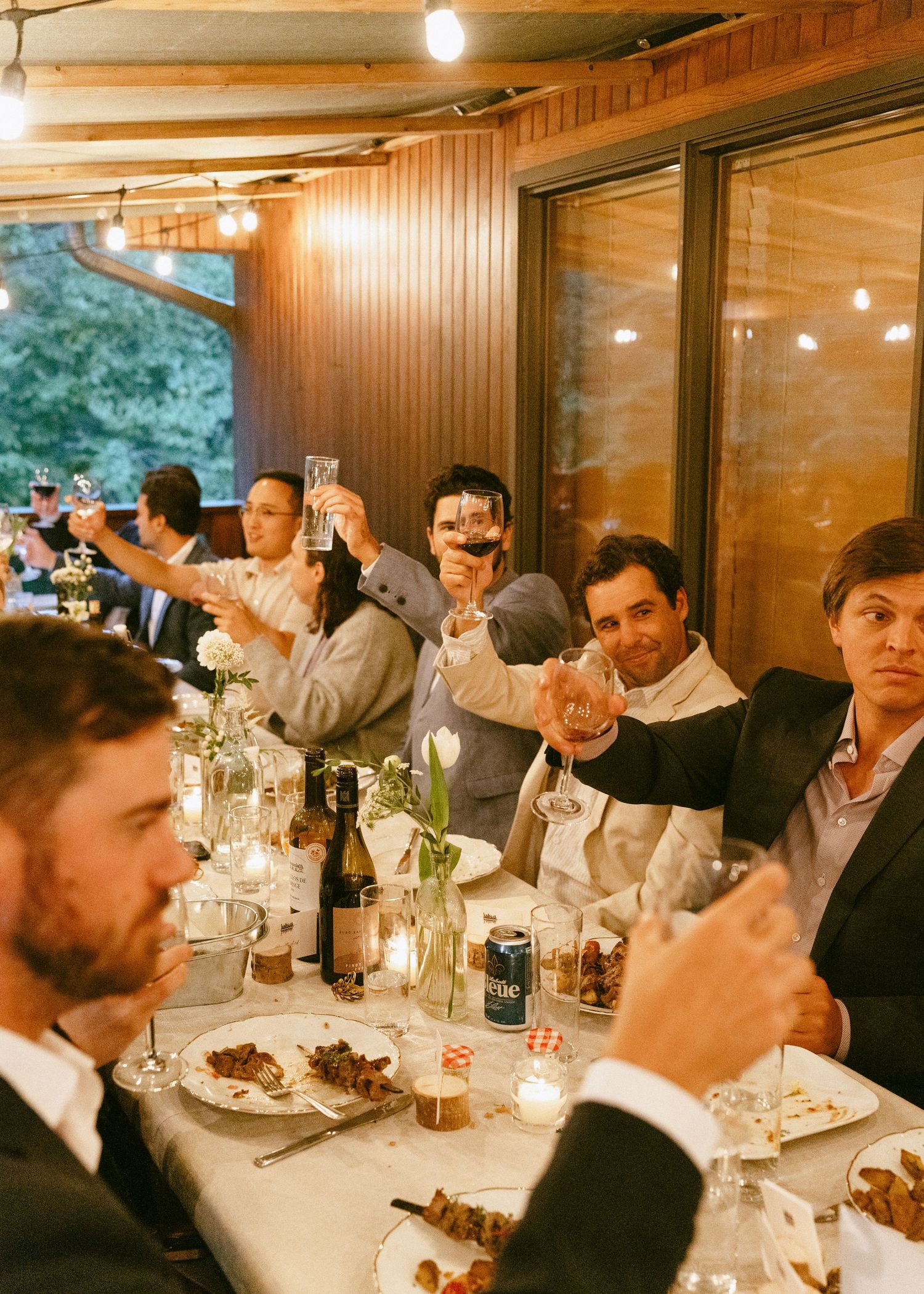 Men raising glasses in a toast at a candlelit wedding reception dinner table with string lights.