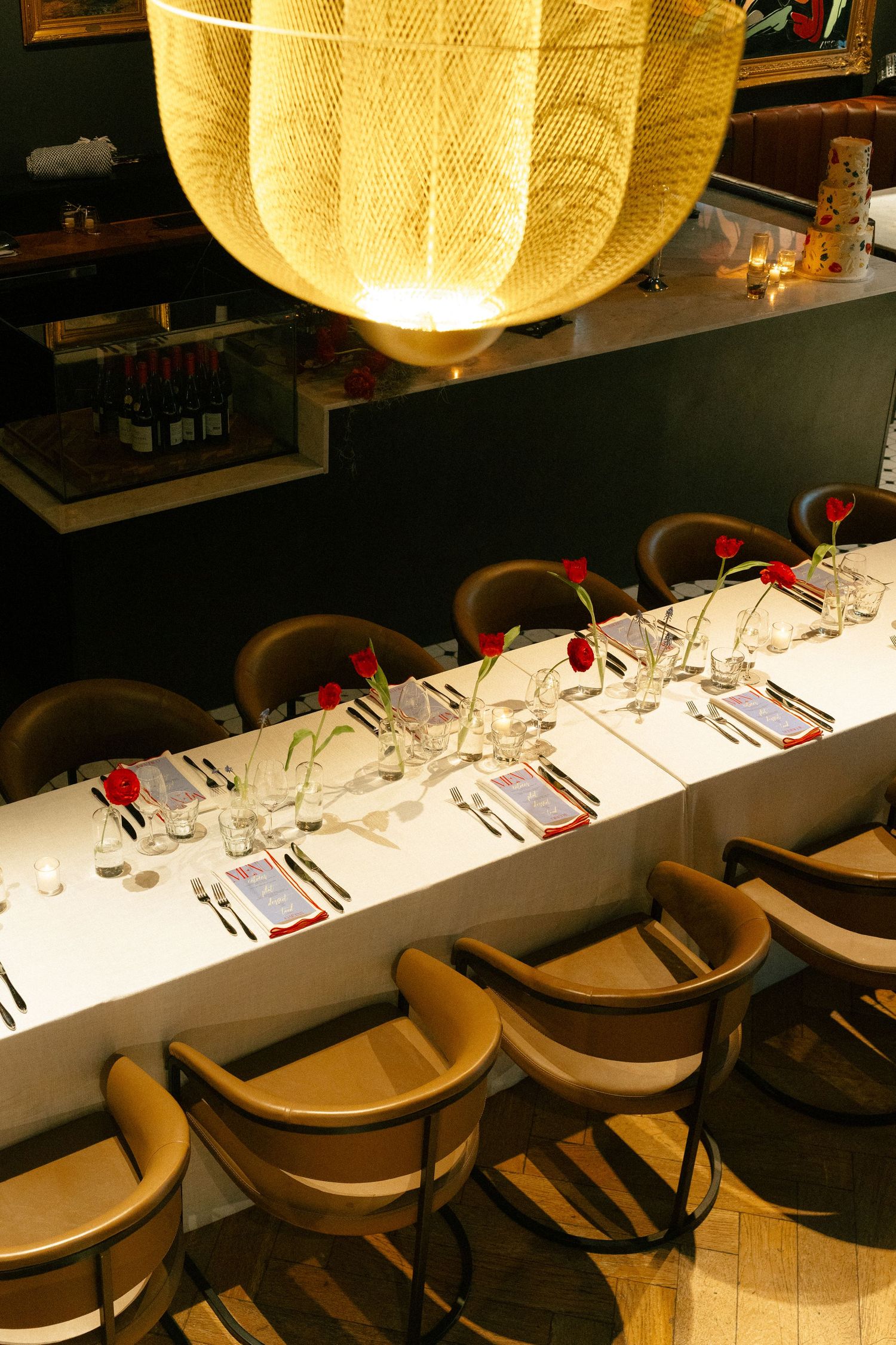 Aerial view of a long banquet table with red tulip centerpieces under a woven pendant light in a dark restaurant.
