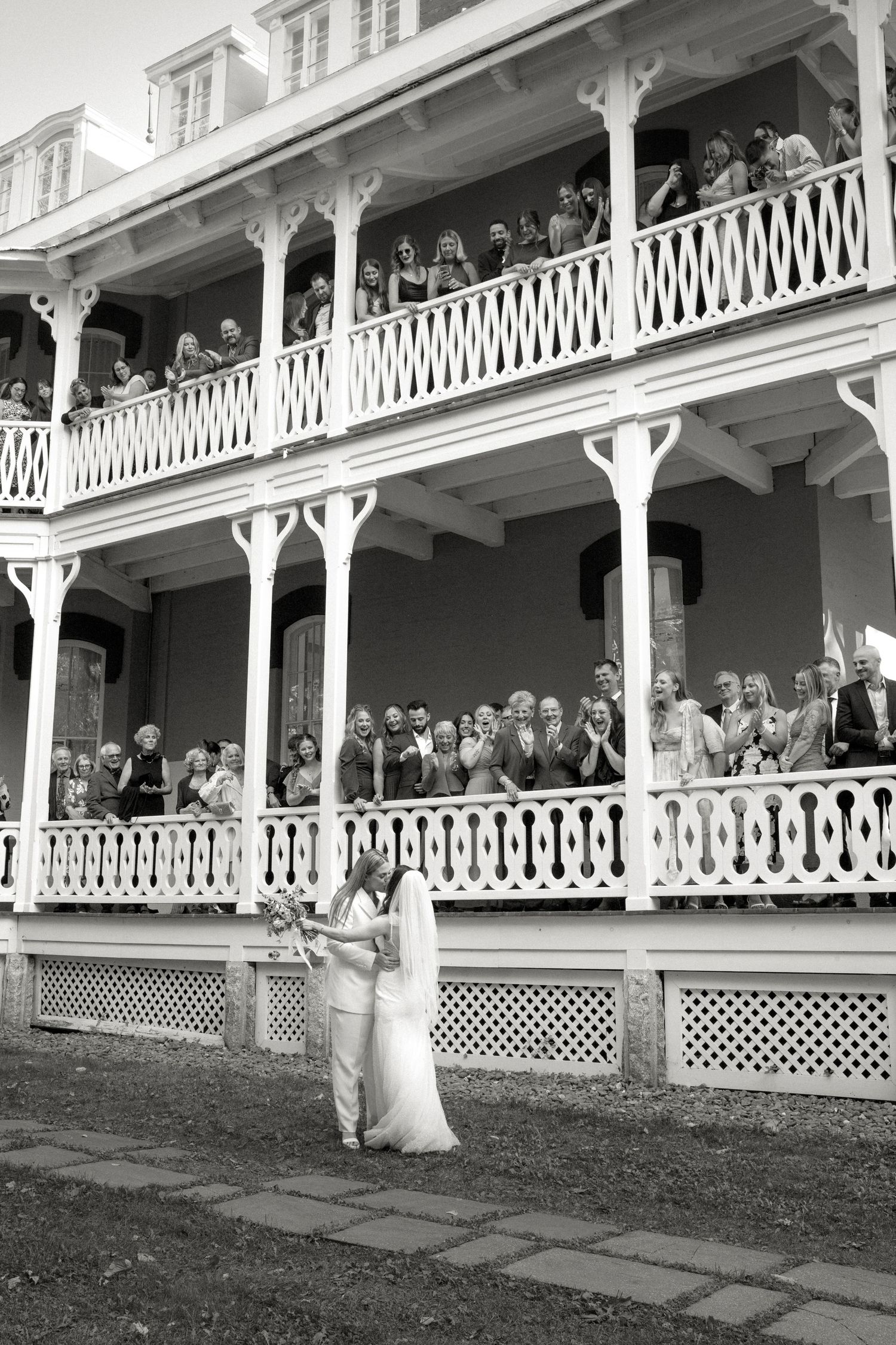 Two brides stand together as wedding guests watch from ornate white balconies of a historic mansion.