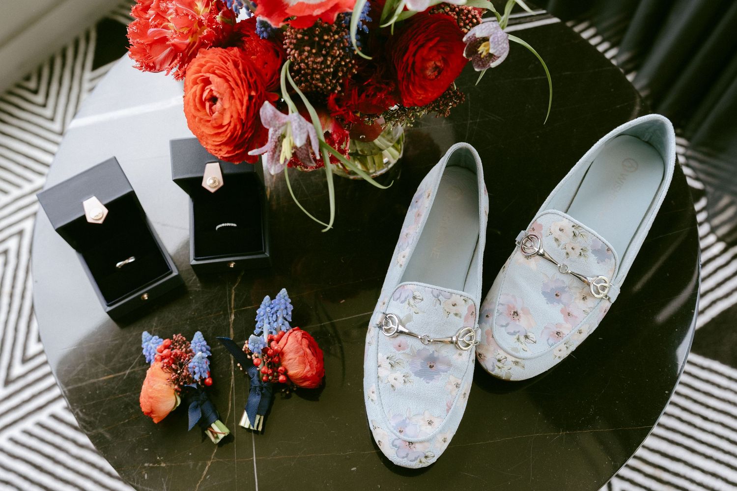 Floral loafers, wedding rings, boutonniere, and red bouquet arranged on dark table as bridal details.