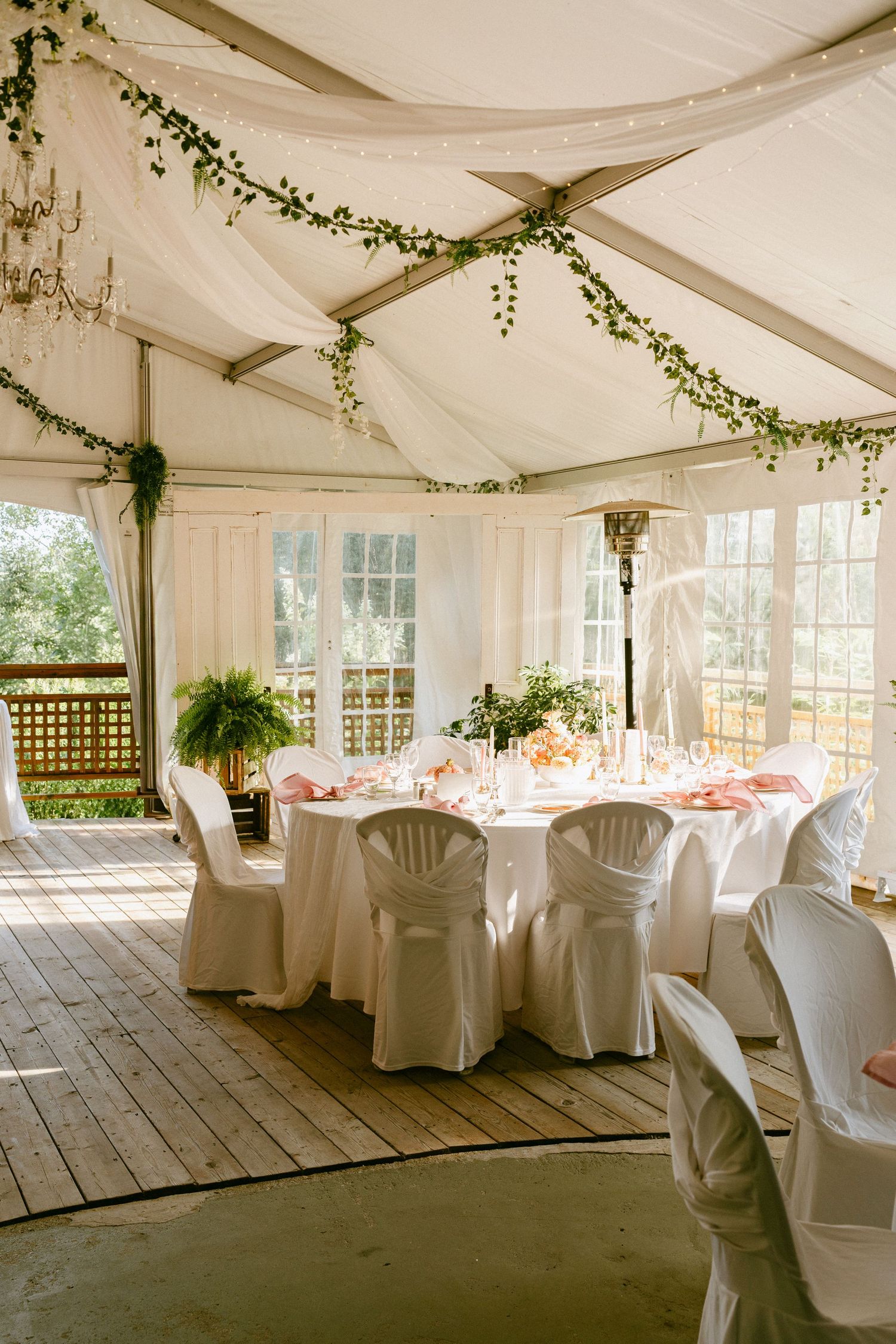 White tent wedding reception with draped ceiling, greenery garlands, round tables, and white chair covers.