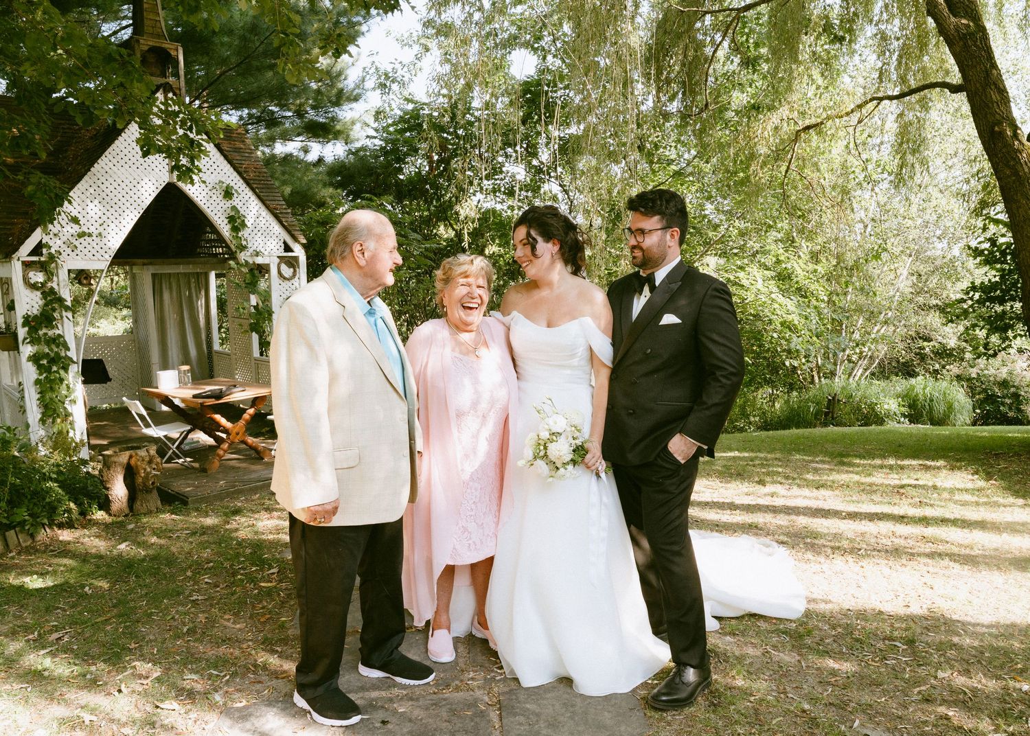 Newlyweds pose with elderly couple in pink outfit at a charming outdoor garden wedding venue.