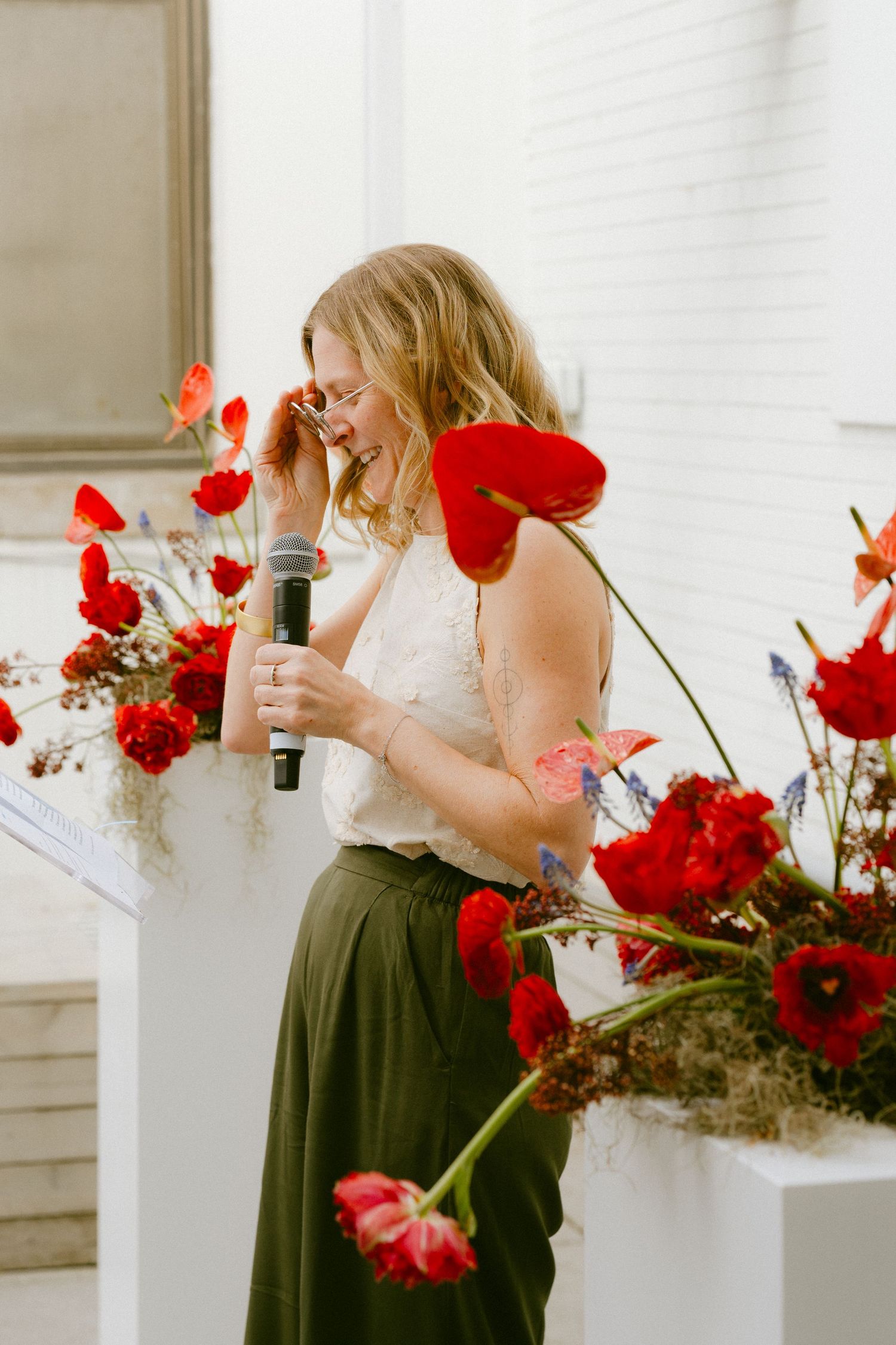 Woman speaking at a podium with a microphone surrounded by lush red poppy floral arrangements at a wedding.