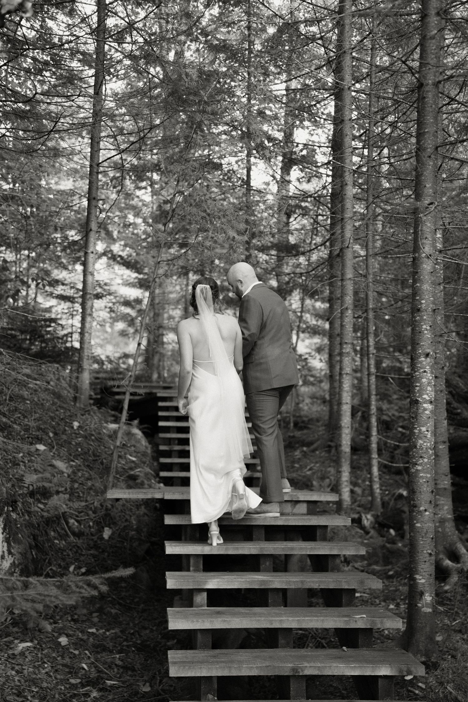 Bride and groom walking up wooden forest stairs in black and white wedding photo.