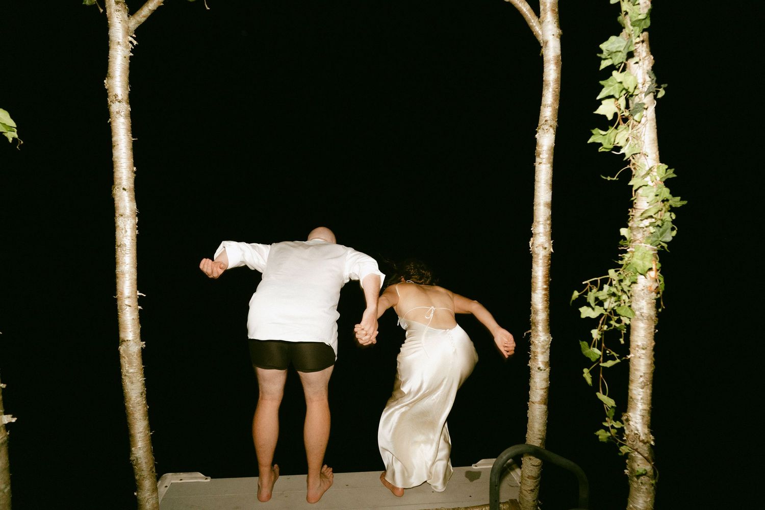Bride and groom jumping together through rustic wooden arch at night.