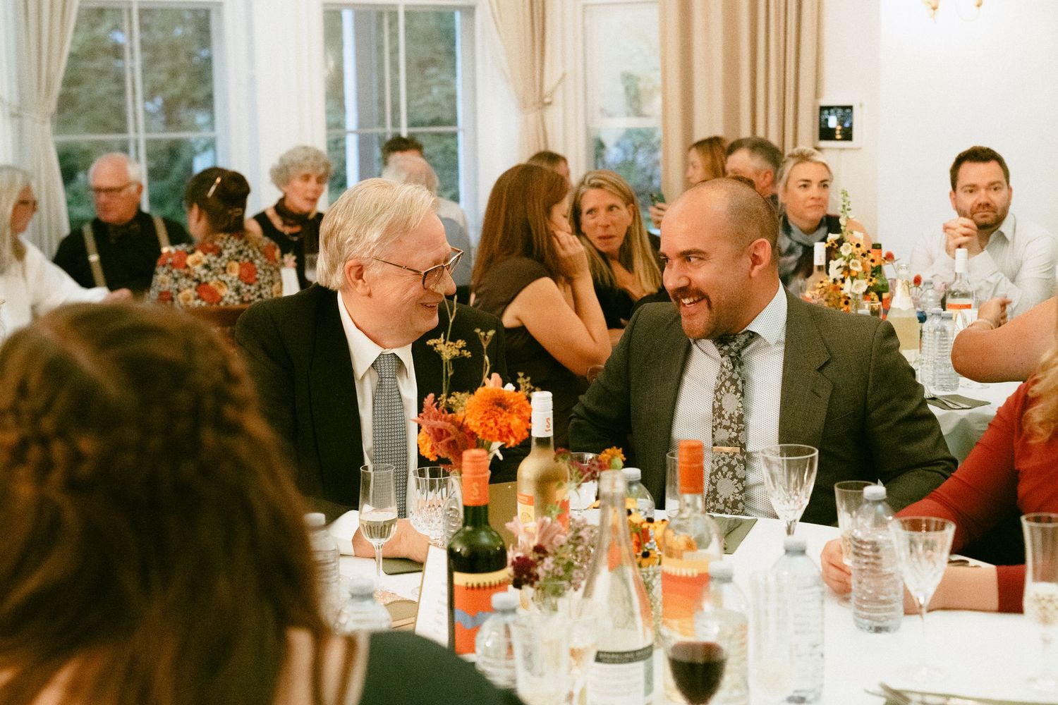 Two men in suits laughing together at a wedding reception dinner table.
