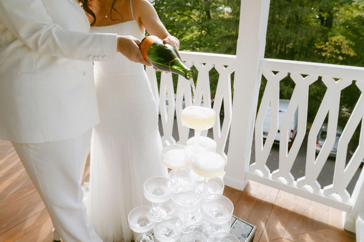 Bride in white gown pours champagne into a tower of glasses at outdoor wedding celebration.
