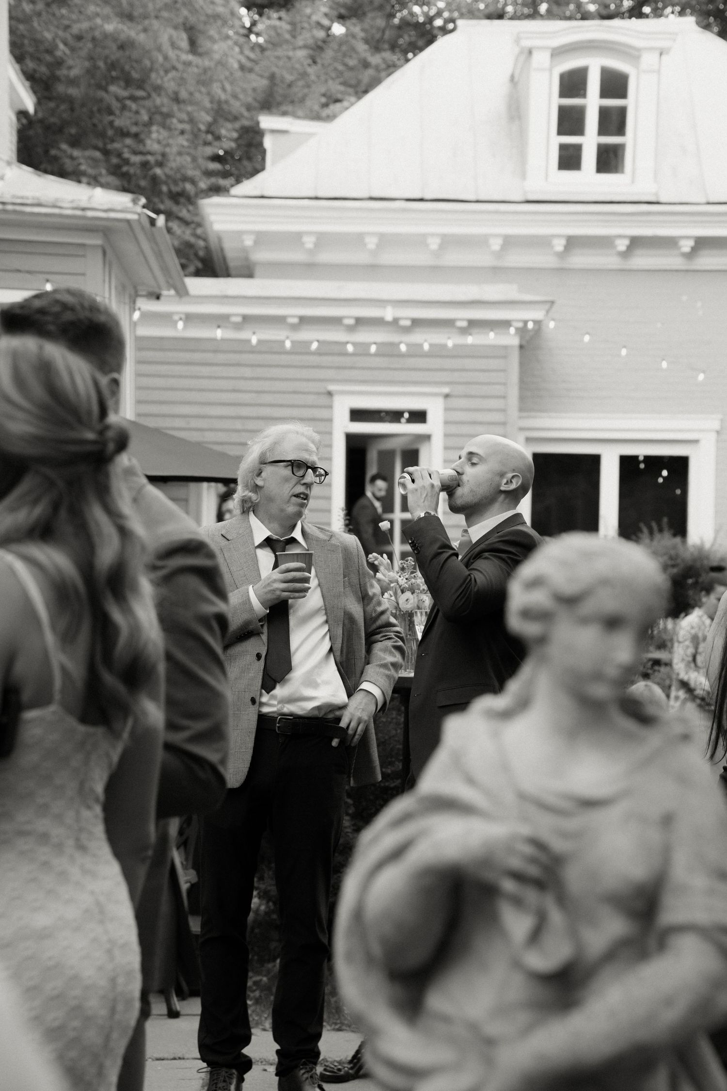 Wedding guests enjoy drinks outdoors near a charming building decorated with string lights at evening reception.