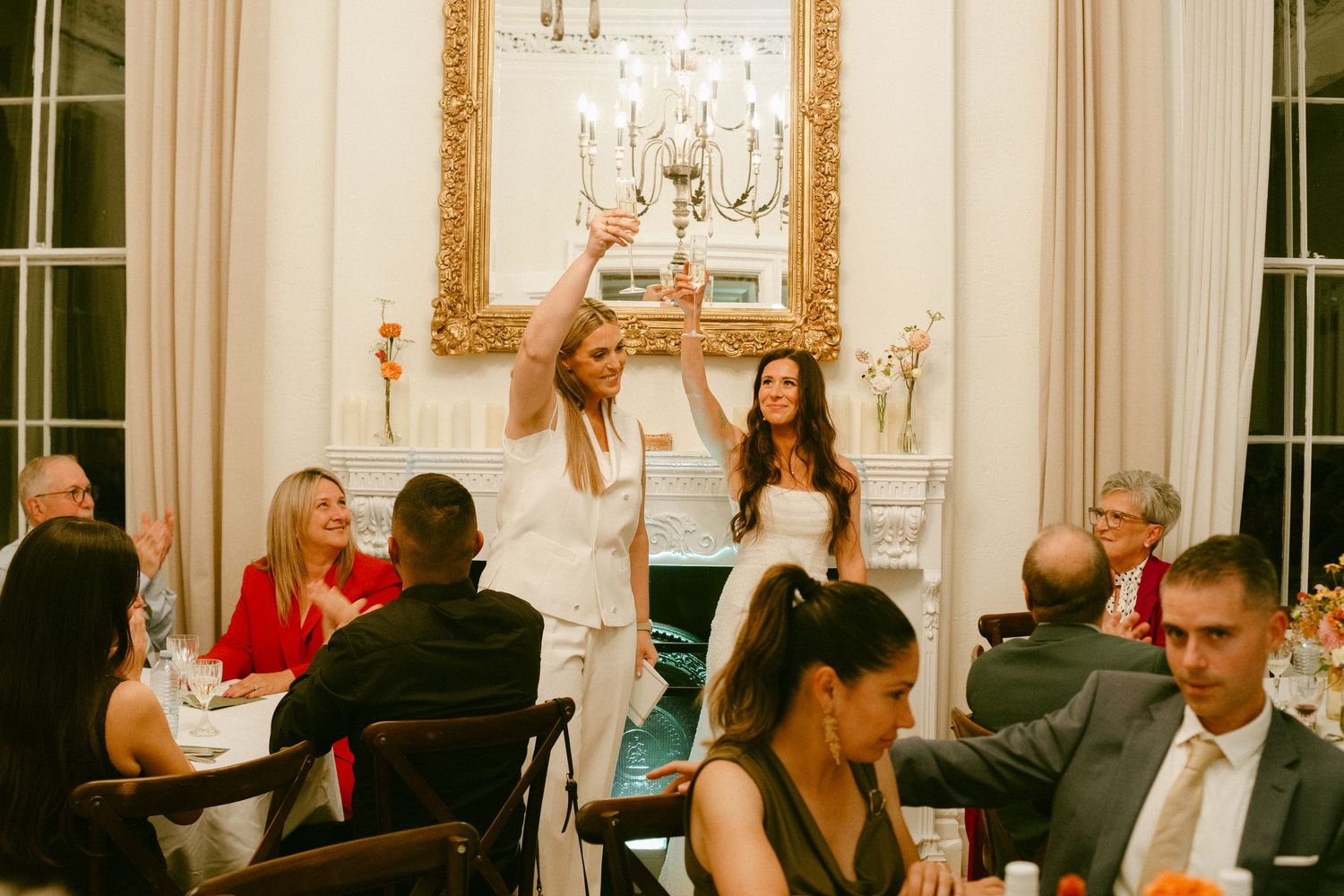 Two brides in white celebrate at elegant wedding reception dinner with guests seated at decorated tables.