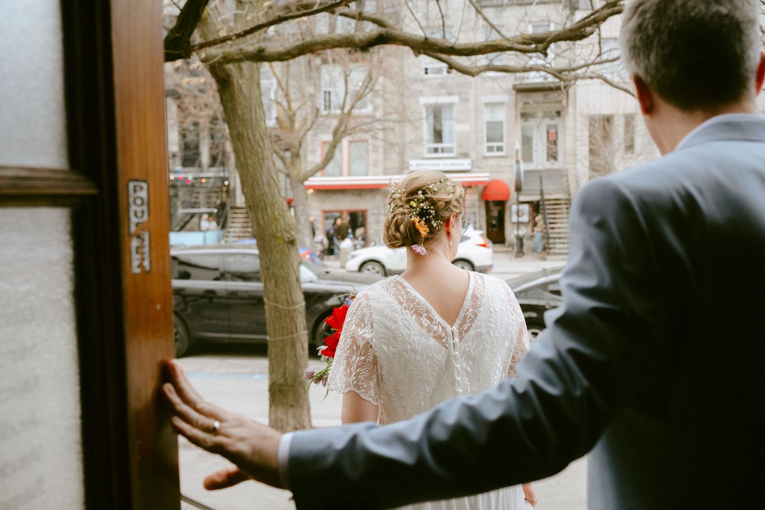 Bride in lace wedding dress with floral updo exits through wooden door onto city street.