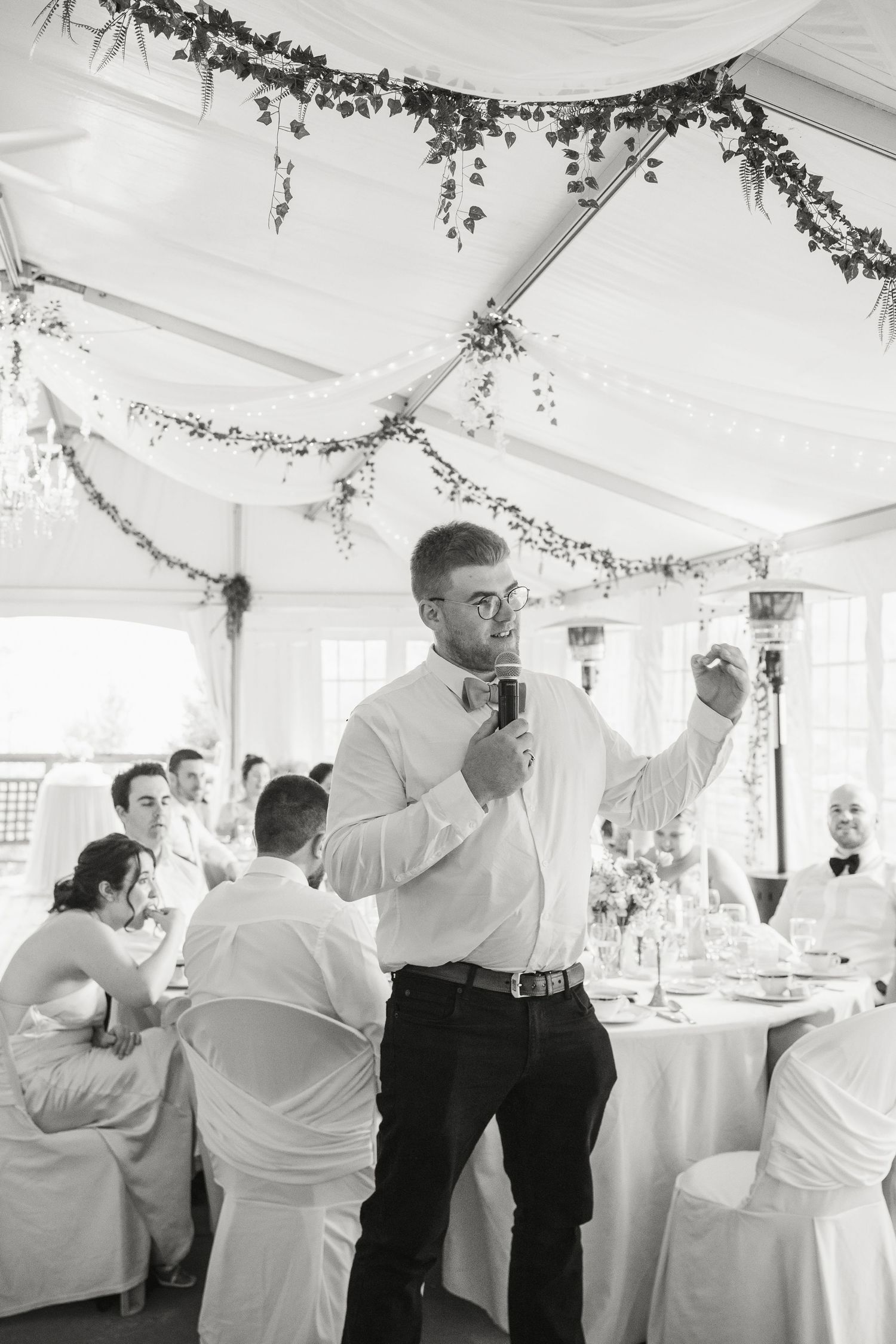 Black and white photo of groomsman giving speech with microphone at wedding reception.