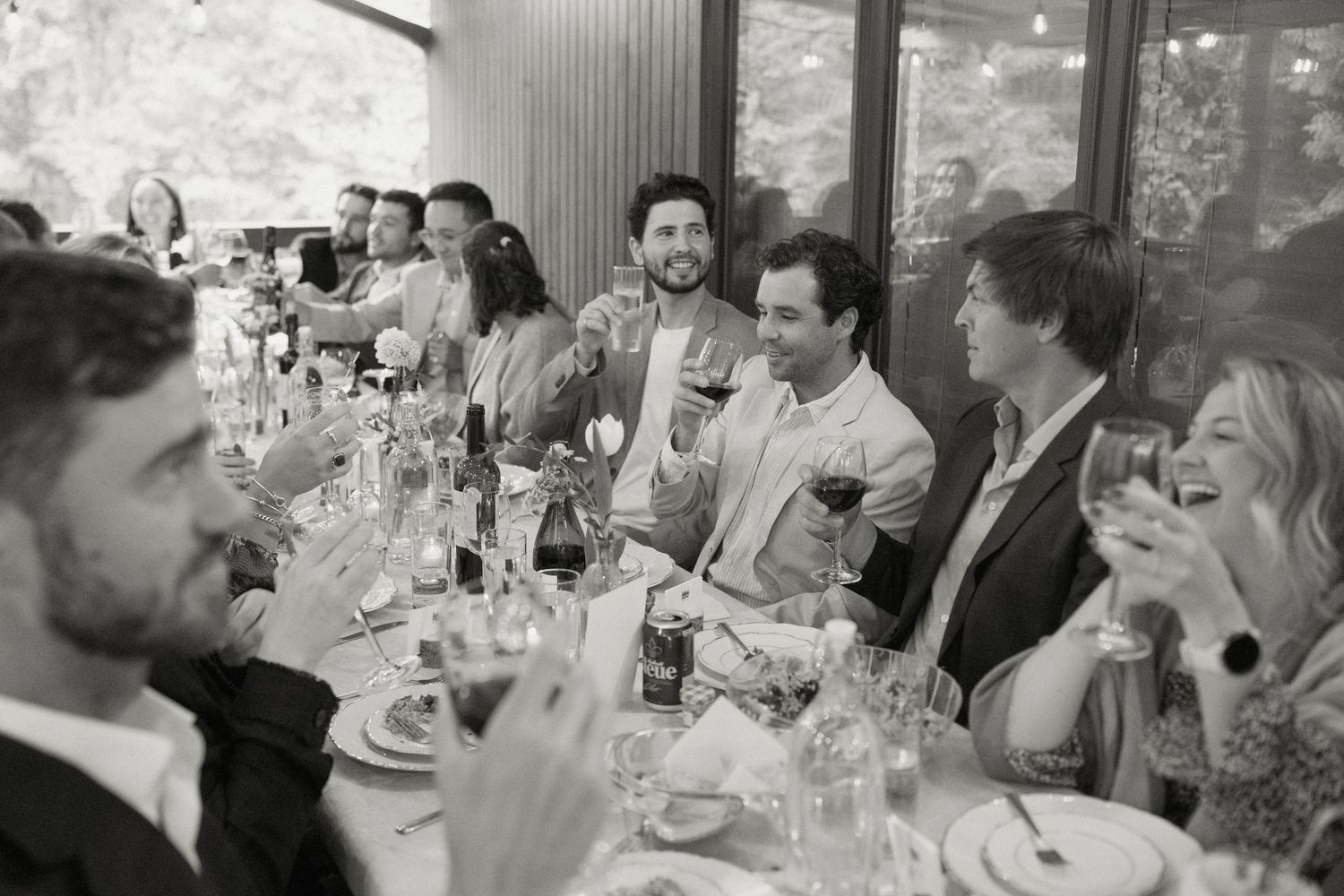 Black and white photo of joyful wedding guests laughing and raising glasses at a reception dinner table.