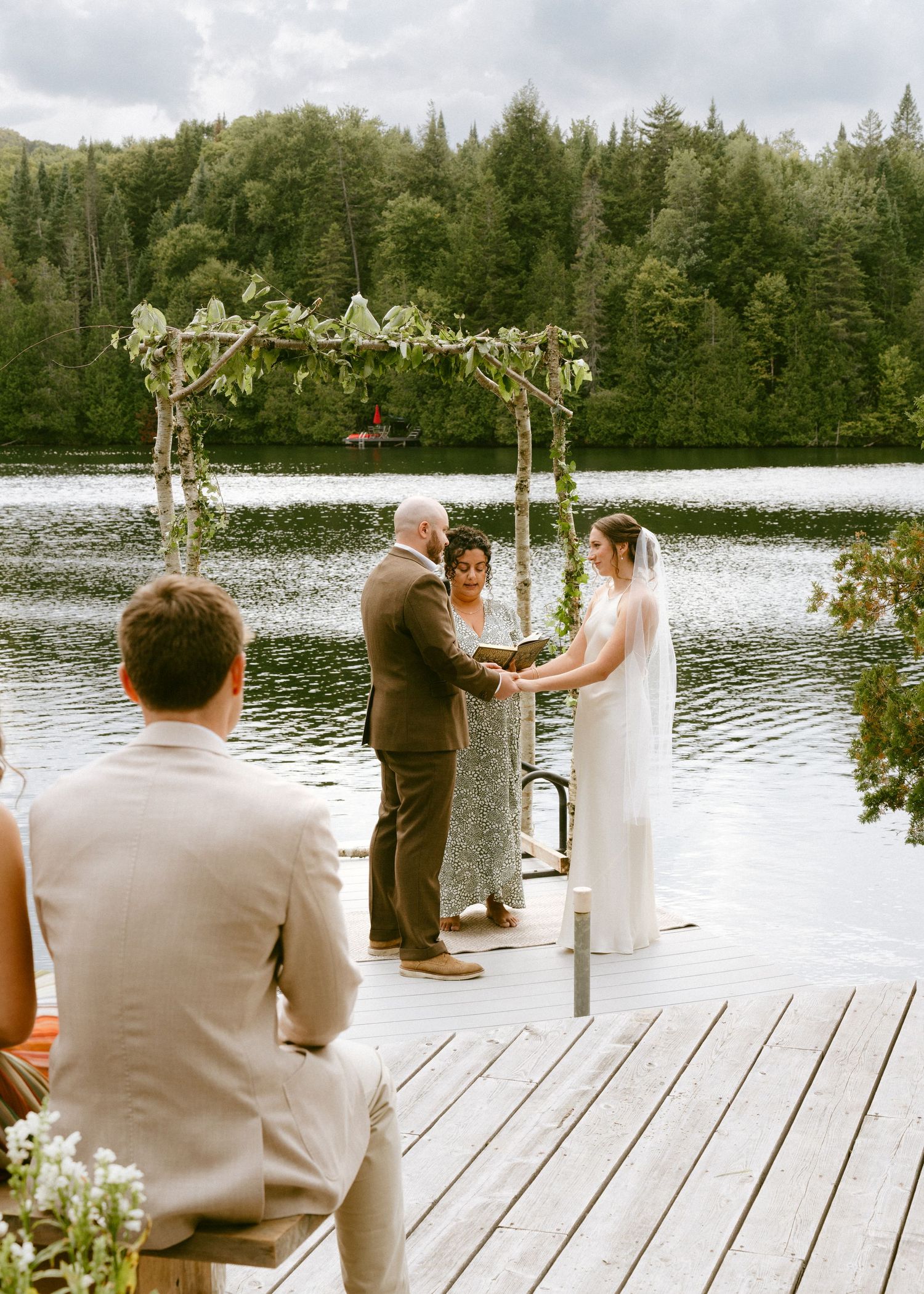 Intimate outdoor wedding ceremony on a lakeside dock with a greenery arch and small guest gathering.