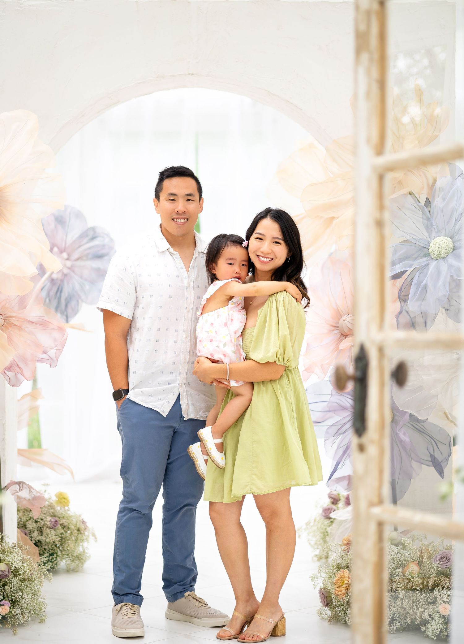 Happy Asian family portrait with parents holding toddler daughter amid pastel floral backdrop.