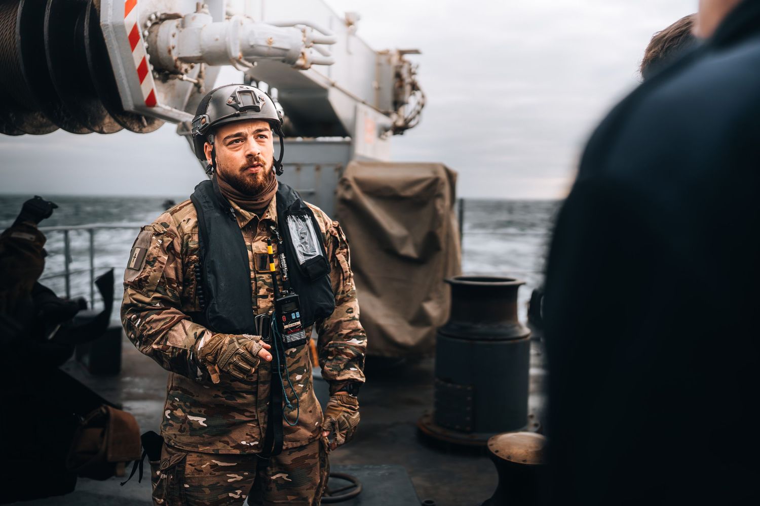 Bearded man in military vest and helmet standing on ship deck at sea.