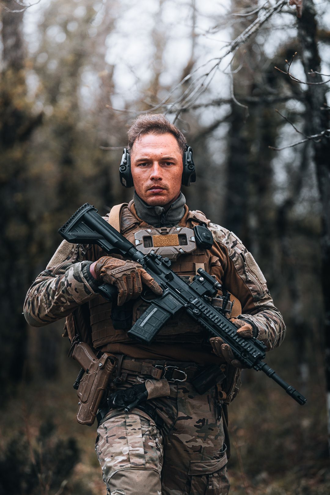 Armed soldier in camouflage gear holding rifle in forest during overcast day.