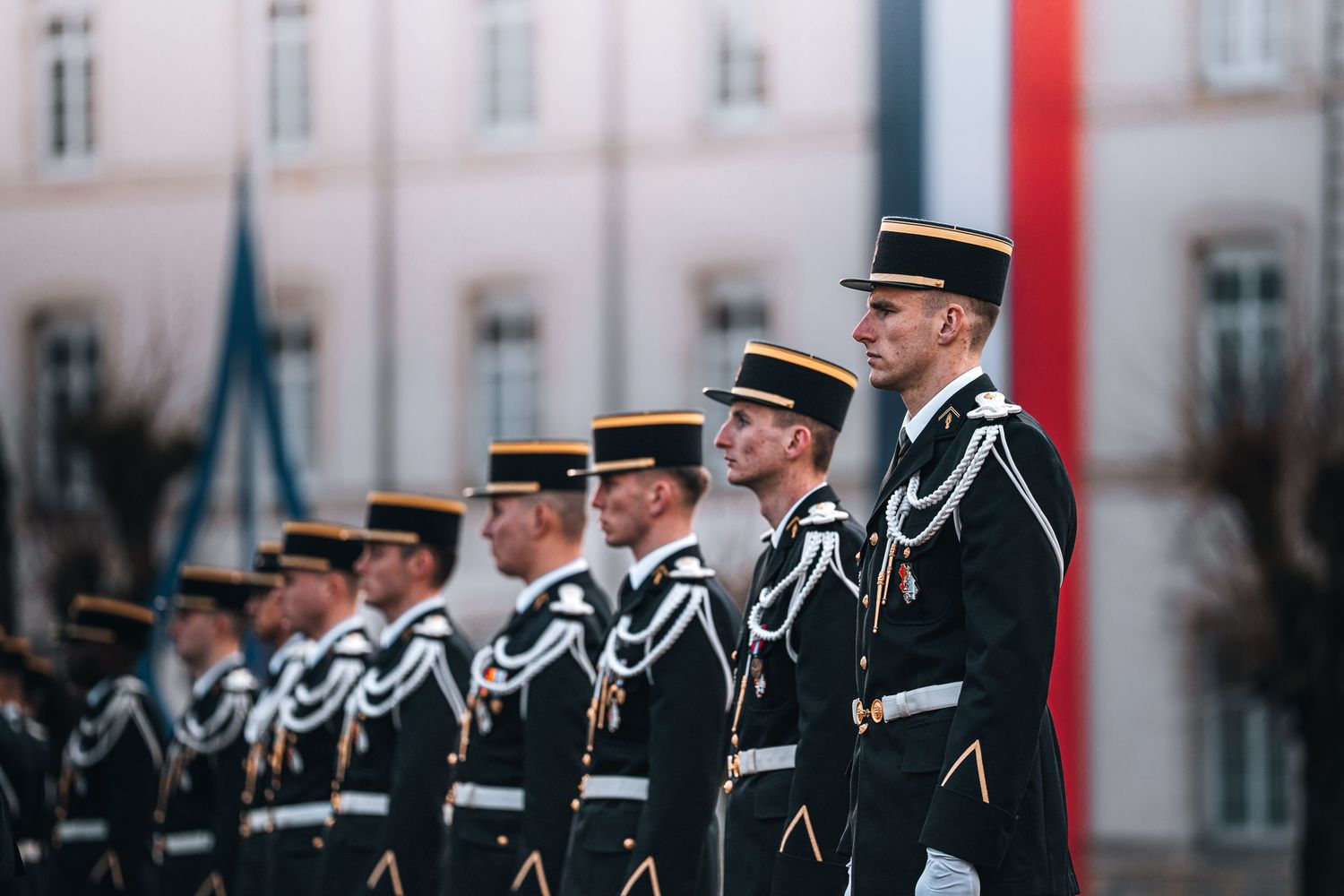 A row of French military officers in formal black uniforms with gold braiding stand at attention during a ceremony.