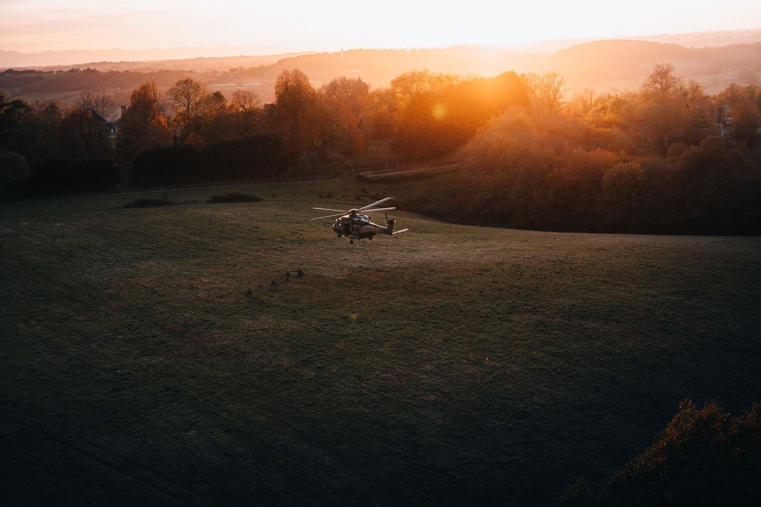 Military helicopter landing on green field at golden sunset with hills.