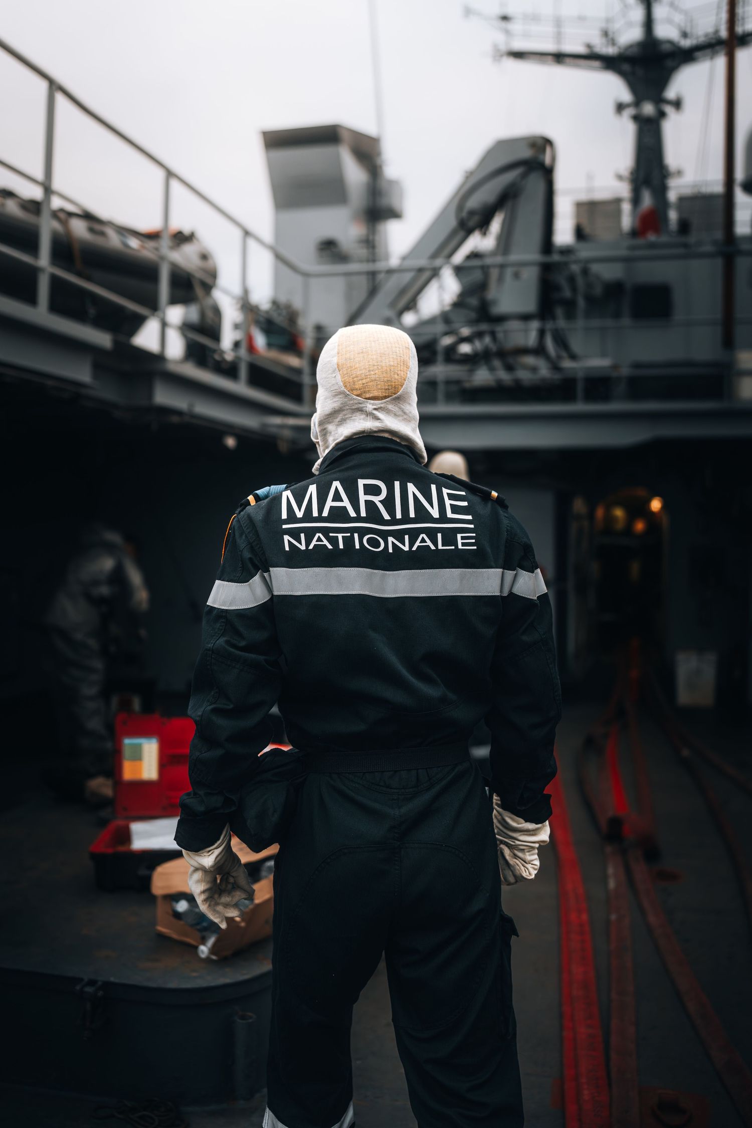 French Marine Nationale sailor stands on warship deck wearing protective hood and uniform.