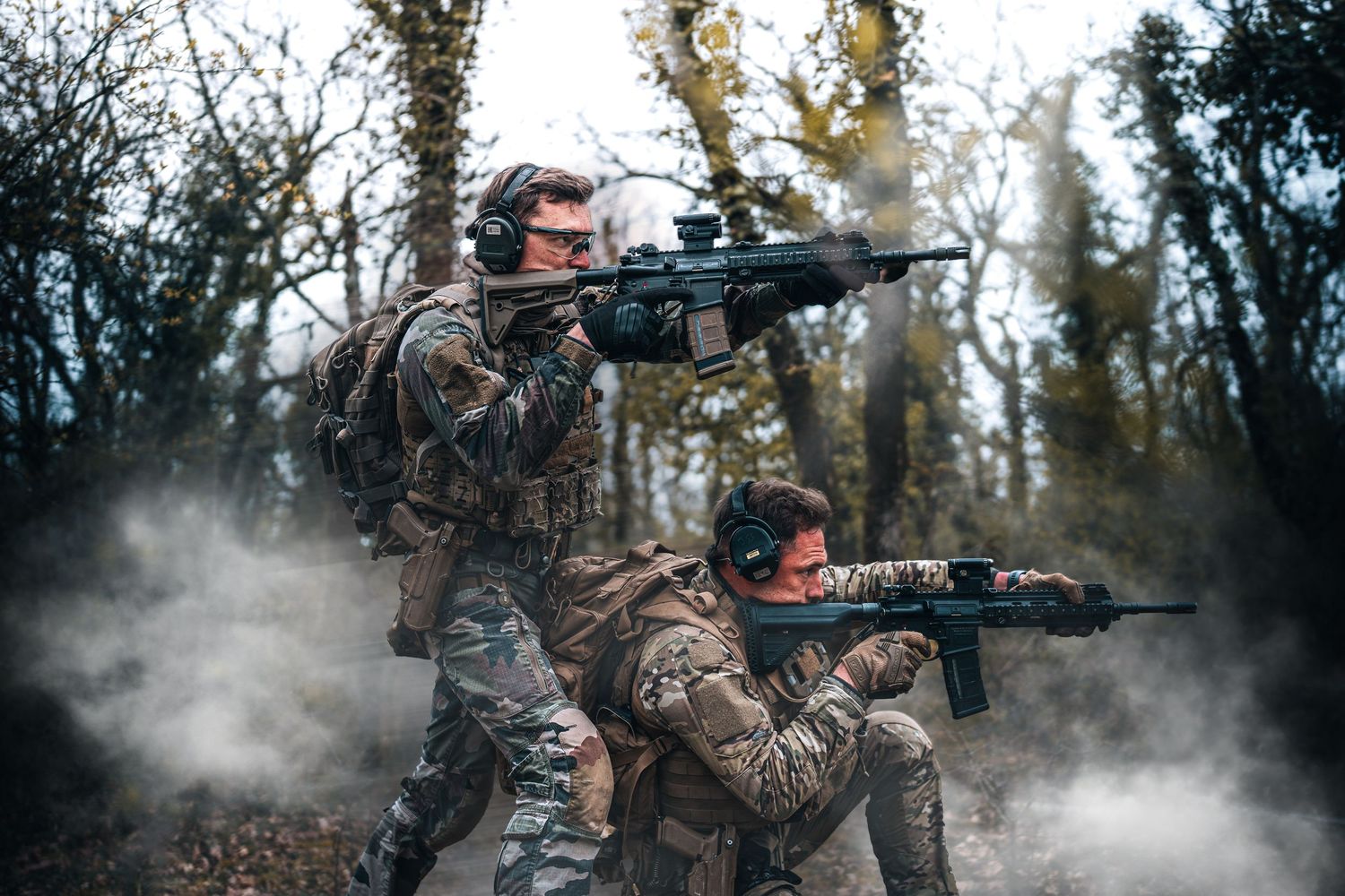 Two soldiers in camouflage aiming rifles in smoky forest during military exercise.