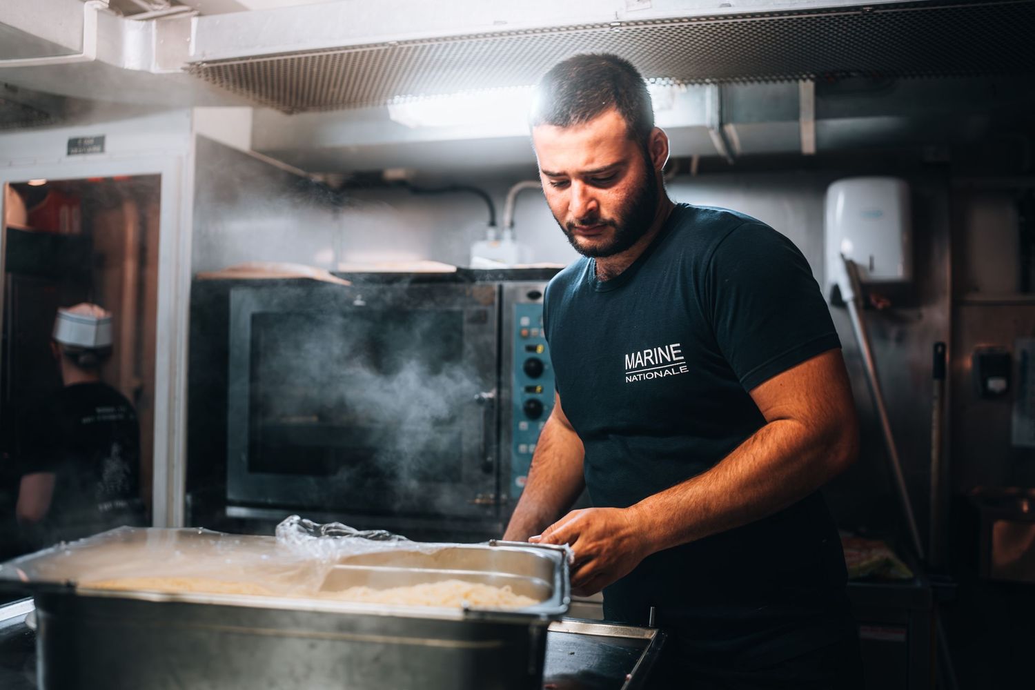 Navy cook in black uniform preparing large batch of food in professional ship kitchen.