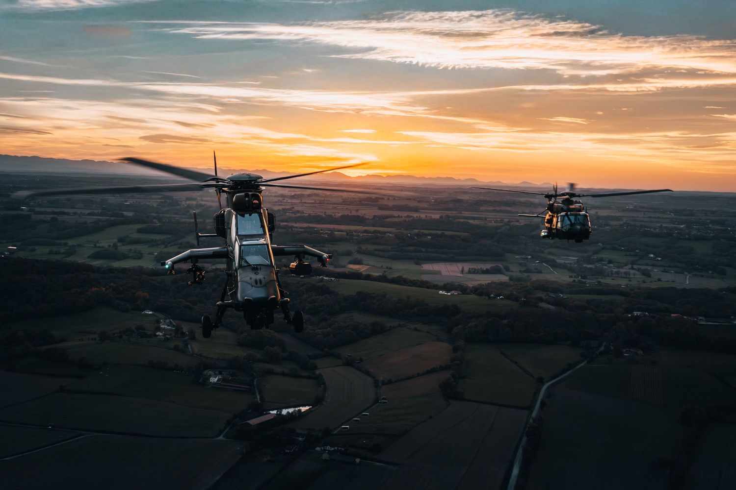 Two military helicopters flying in formation over green fields at sunset with vibrant orange and purple sky.