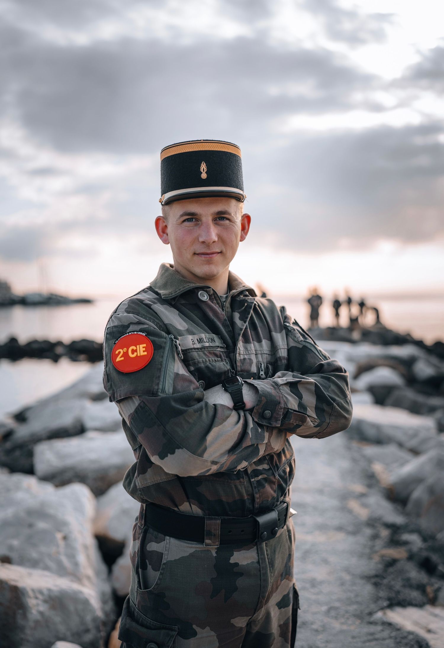 French soldier in camouflage uniform and kepi hat stands with arms crossed near rocky shoreline at sunset.