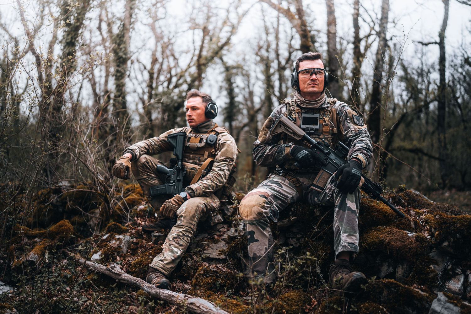 Two camouflaged soldiers rest on rocks in forest, one holding rifle during outdoor military training.