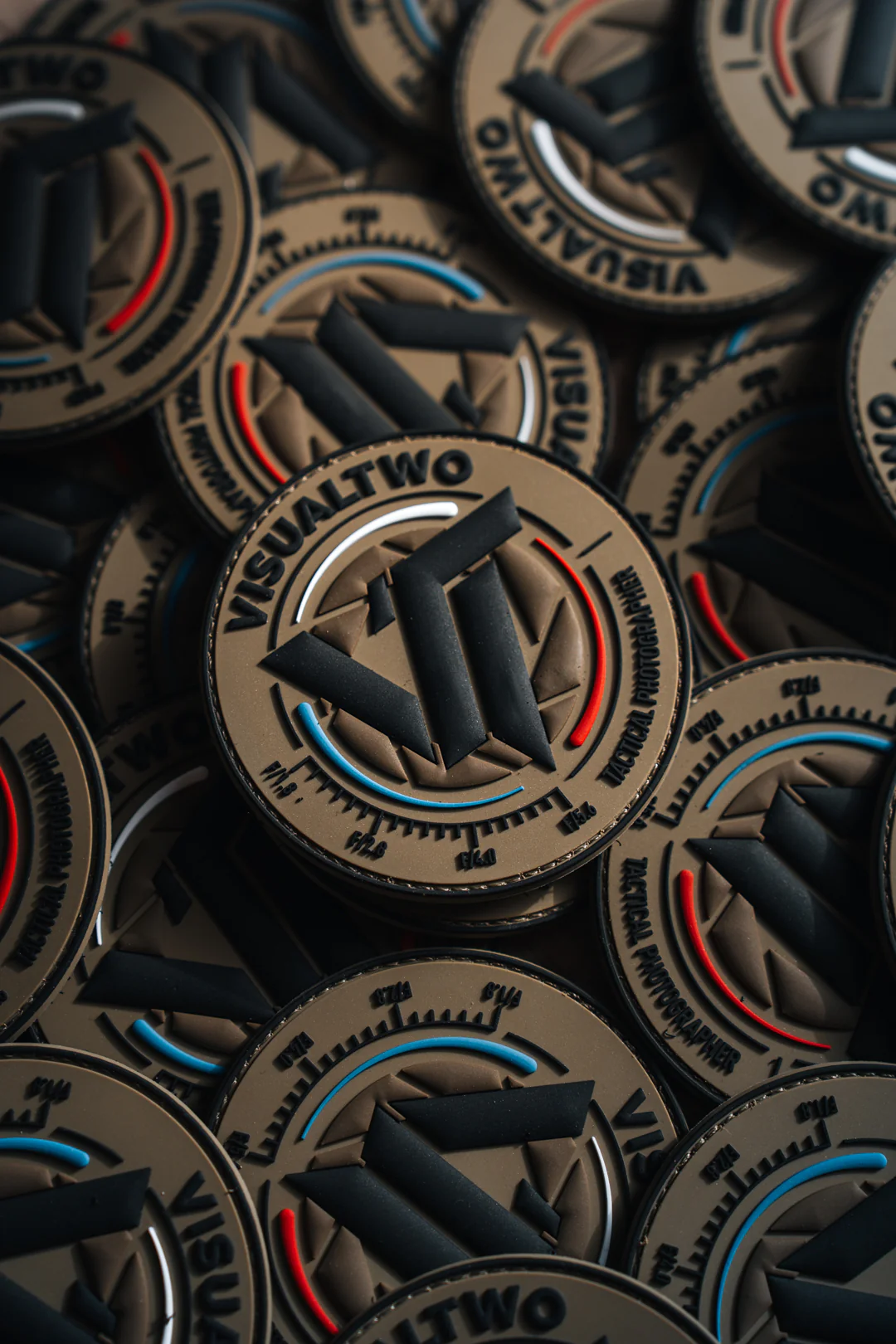 Pile of round VISUALTWO branded PVC patches with black logo design, red and blue accent lines on tan background.