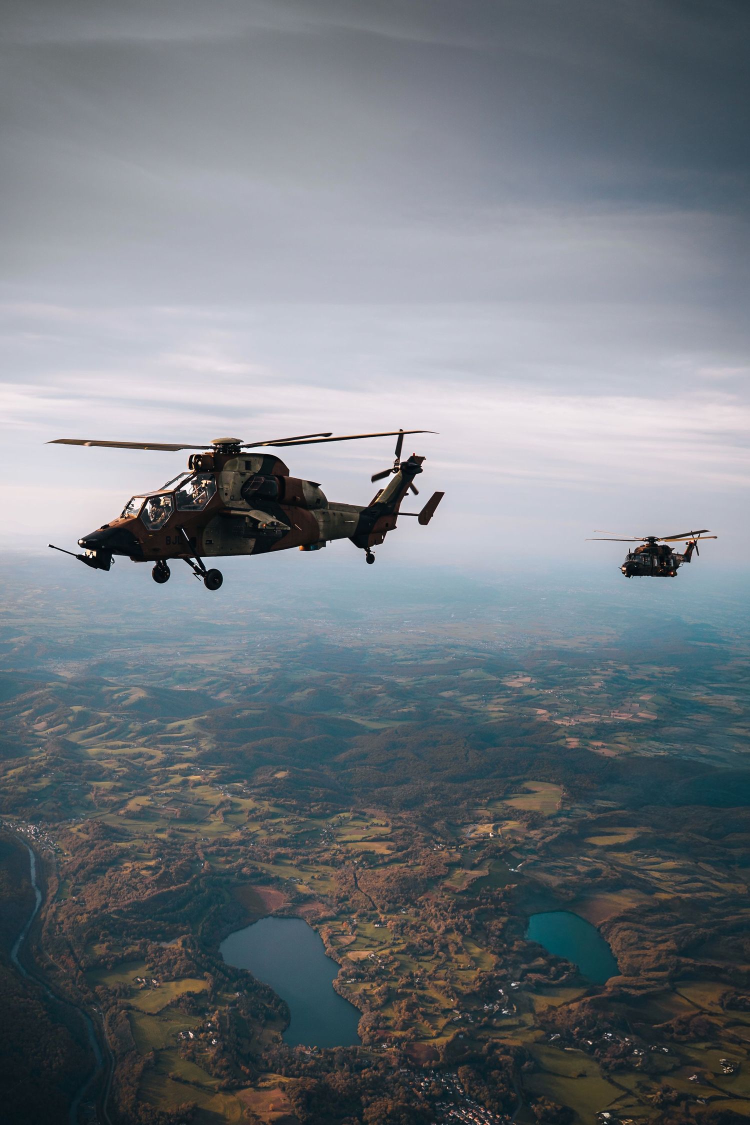 Two military helicopters flying over scenic landscape with lakes and fields below.