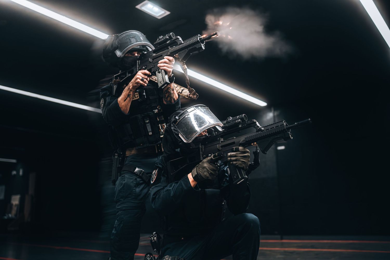 Two tactical operators in black gear fire rifles in low-light indoor shooting range with smoke.