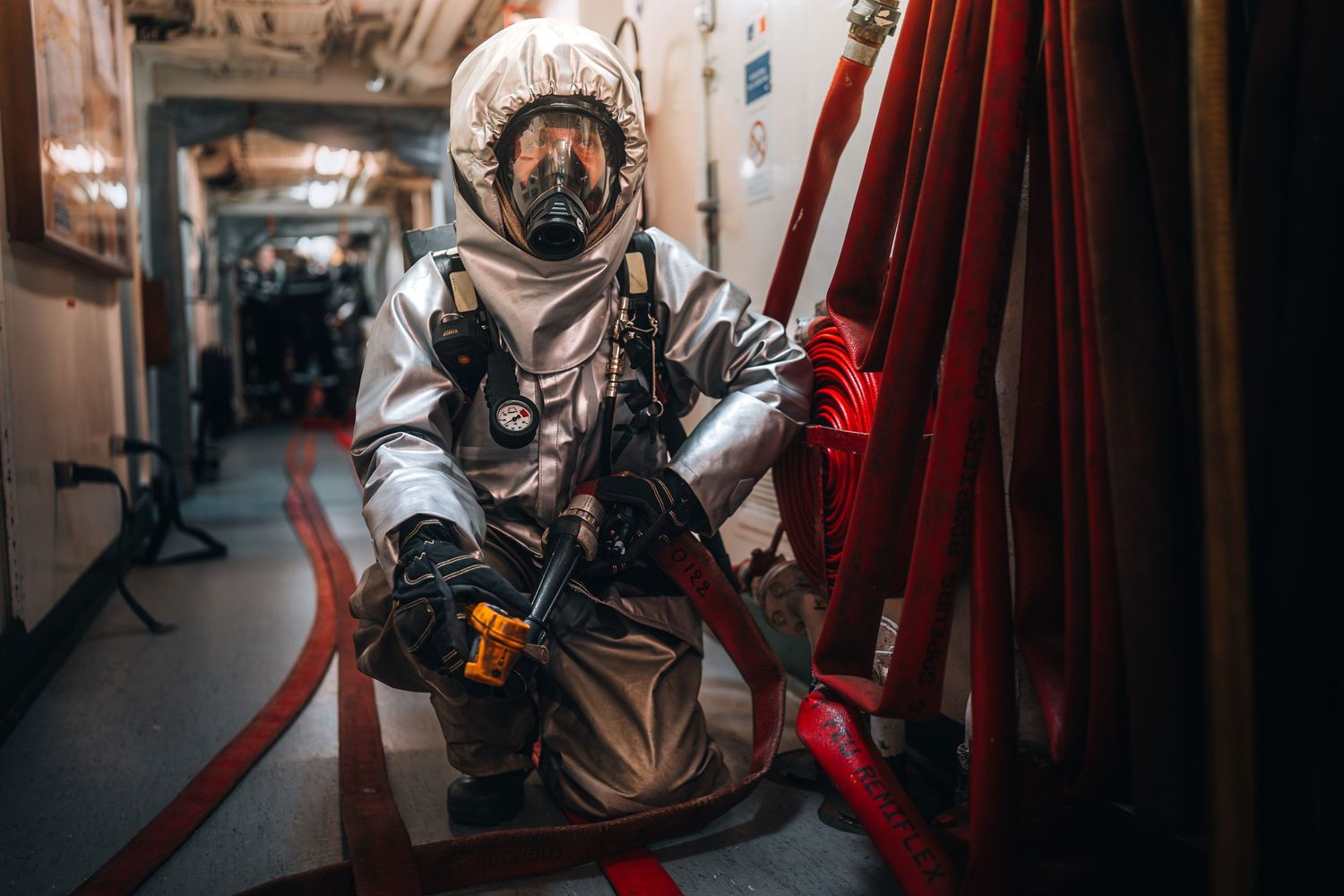 Worker in full silver hazmat suit and gas mask crouching near red hoses.