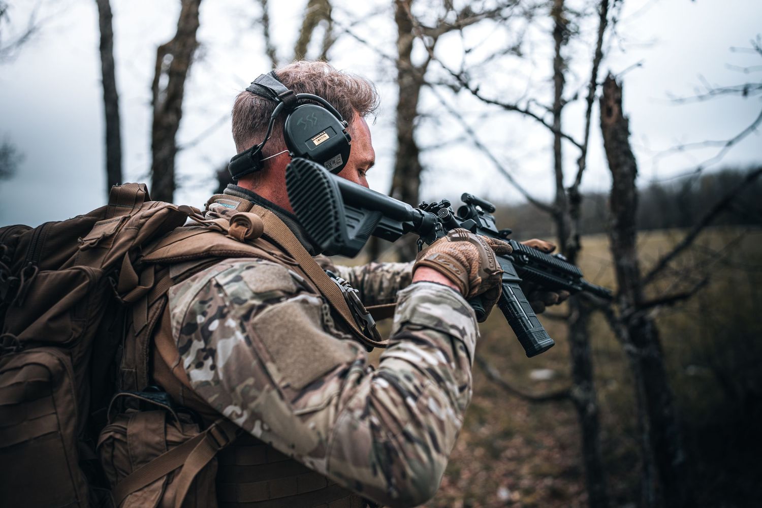 Soldier in camouflage with backpack aiming rifle in bare winter forest.