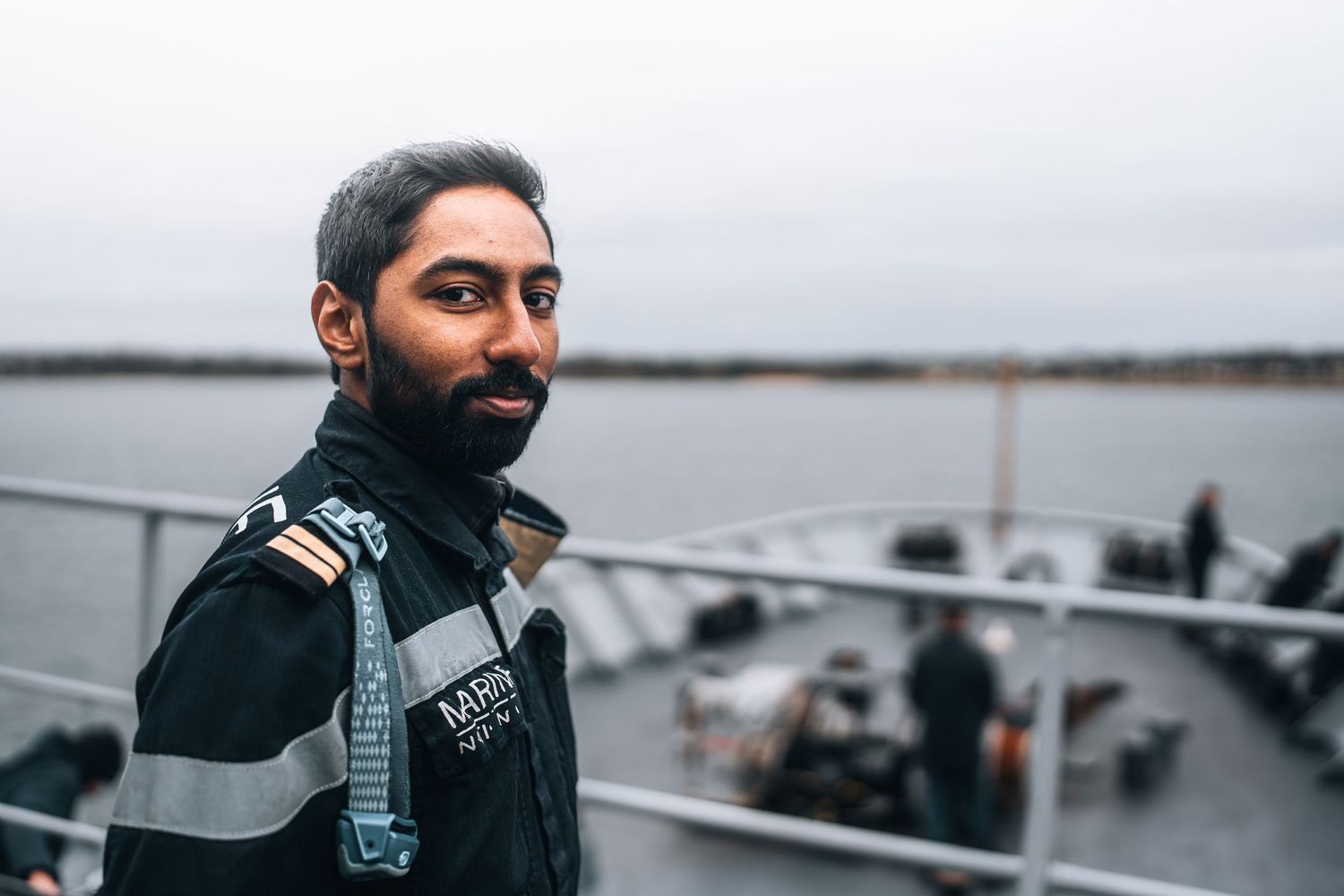 Bearded naval officer in uniform standing on ship deck with water in background.