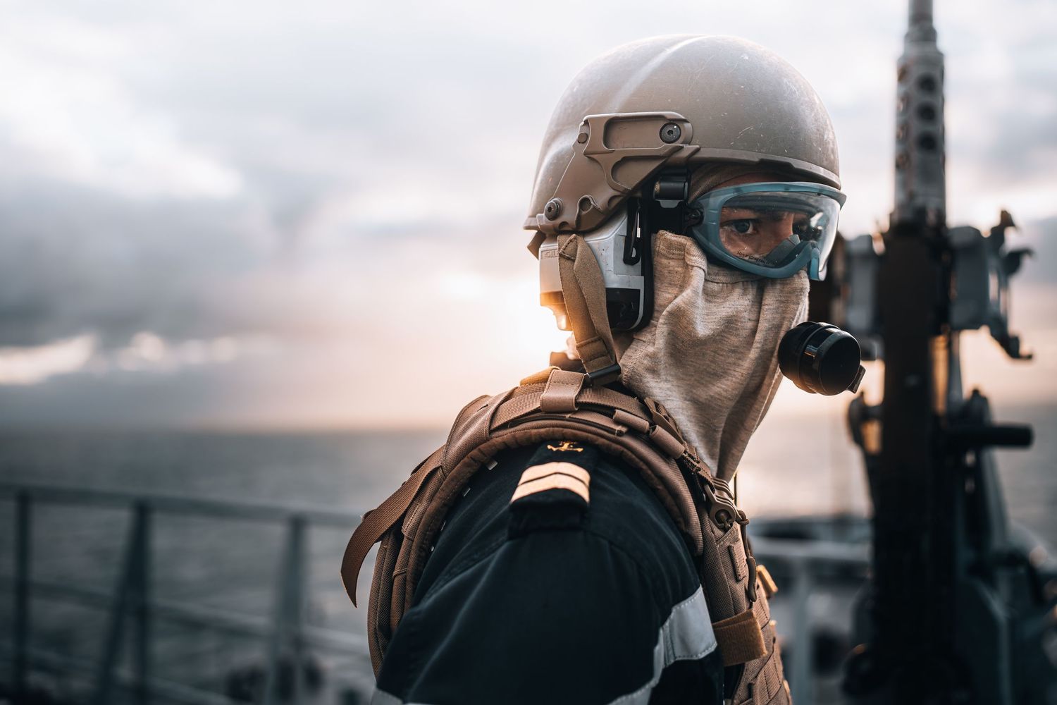 Armed soldier in tactical gear, helmet, and goggles stands on a naval vessel deck at dusk.