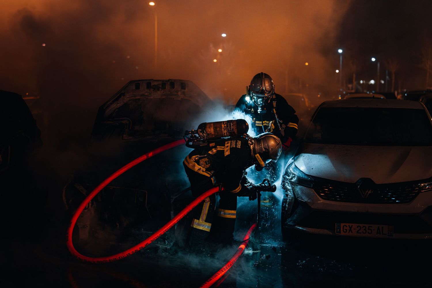 Firefighter battling car fire at night amid smoke and flames in a parking lot.