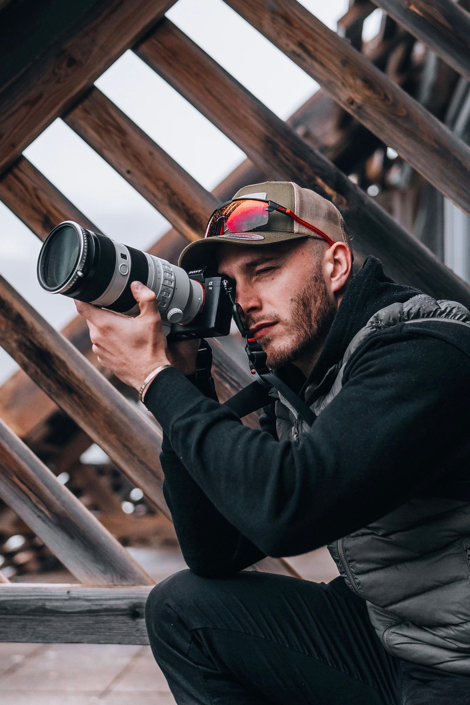 Young male photographer crouching with professional camera and telephoto lens under wooden structure.