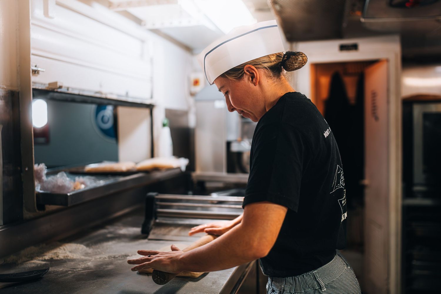 Baker in white hat kneading dough on metal counter in professional kitchen.