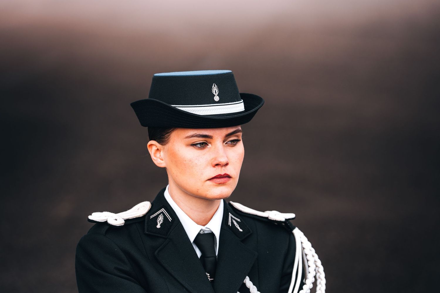 Female military officer in formal black uniform with white epaulettes and decorative hat posing outdoors.