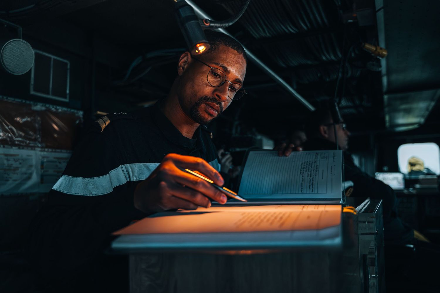 Navy serviceman writing in logbook under dramatic warm lighting inside a military vessel at sea.