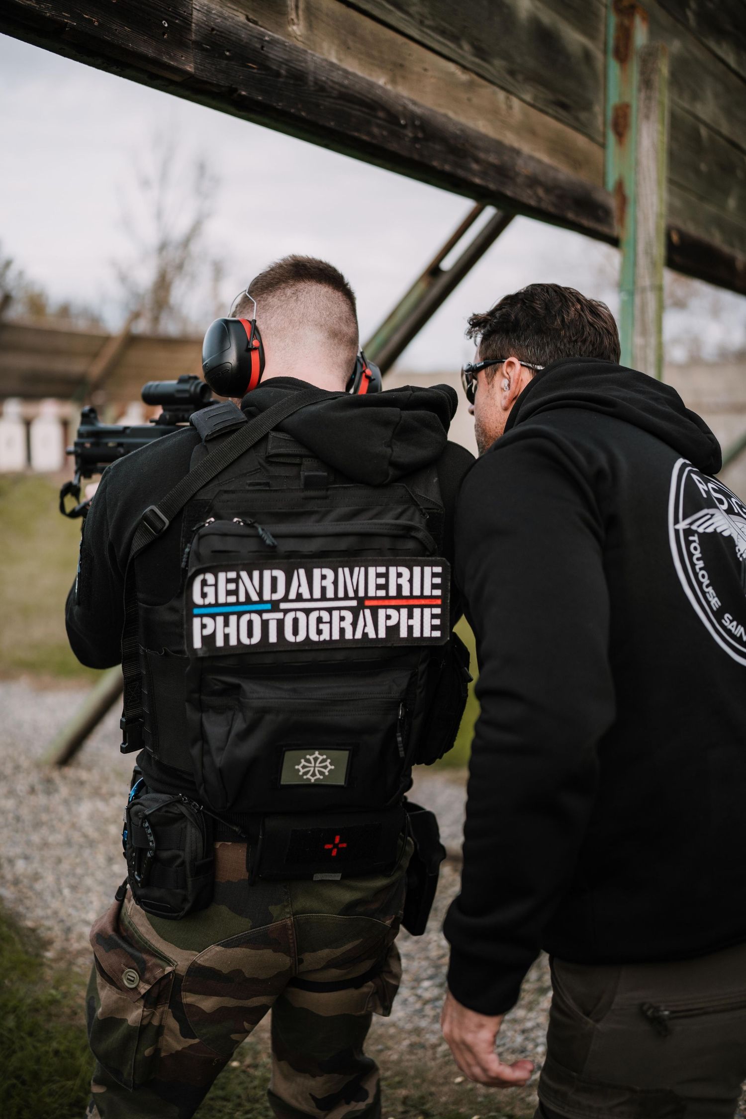 Gendarmerie photographer in tactical gear with camera at outdoor shooting range with instructor nearby.