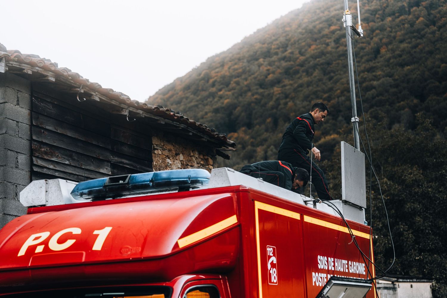 Firefighter standing on top of red PC1 Haute-Garonne fire truck in mountain setting.