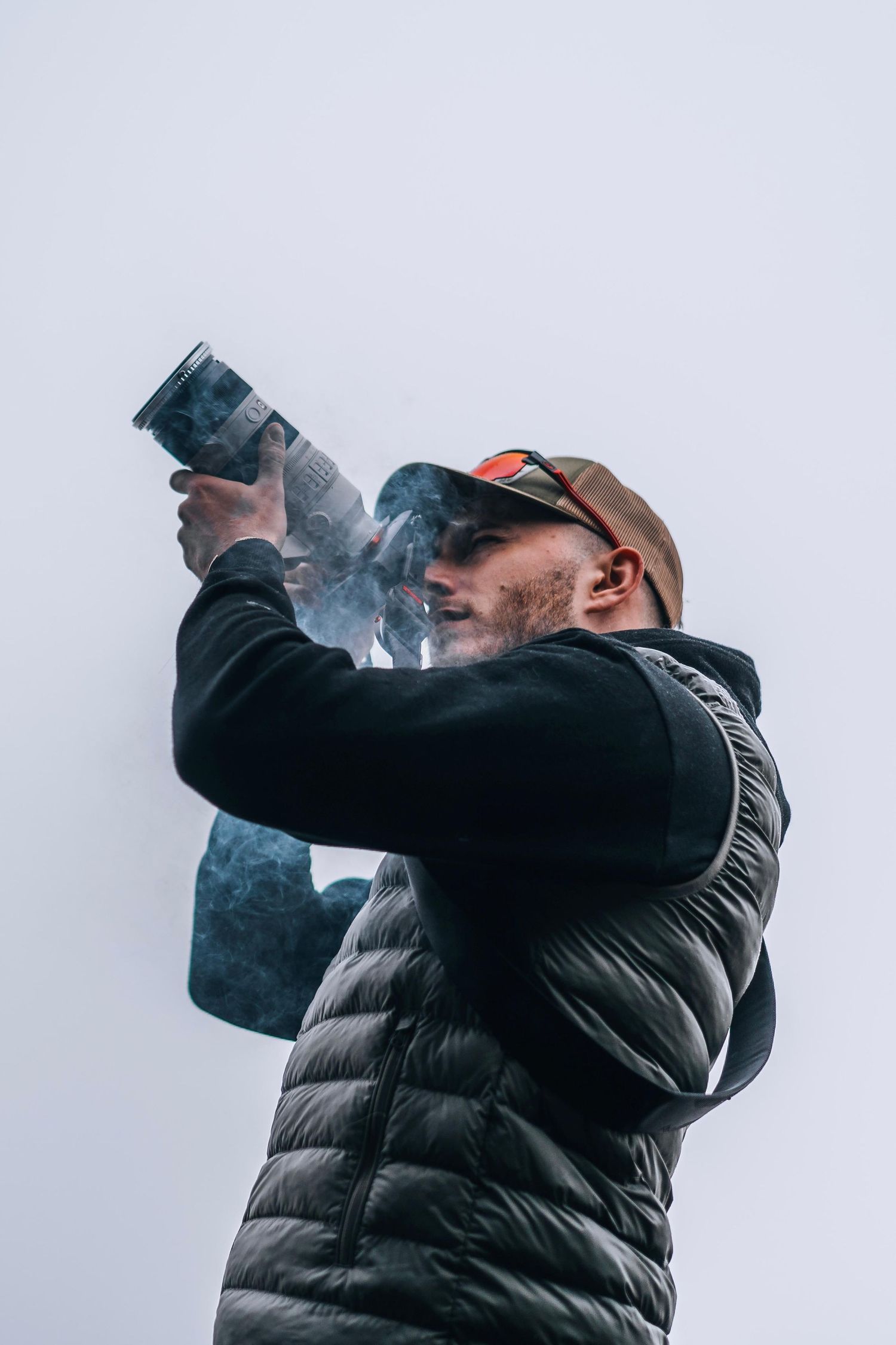 Photographer in a black puffer vest and cap aiming a large camera lens upward against a grey sky.