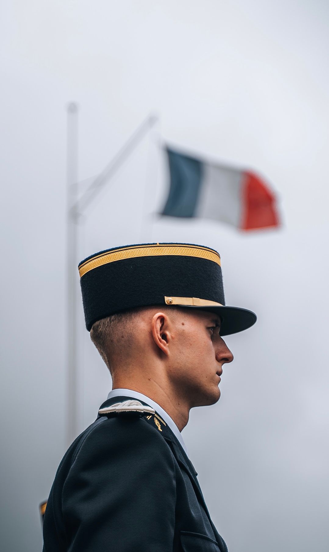 French military officer in dress uniform and kepi hat stands at attention with the French tricolor flag in the background.