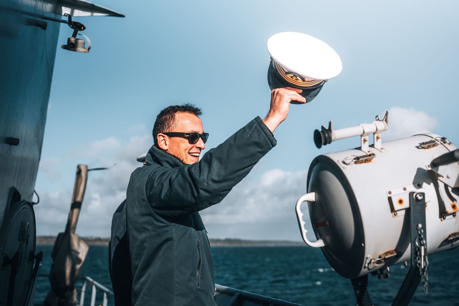 Naval officer in uniform raises white cap while standing on ship deck under blue sky.