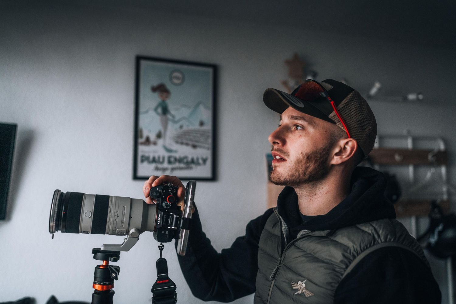 Photographer holding a large telephoto lens camera on a tripod indoors with a framed poster on the wall.