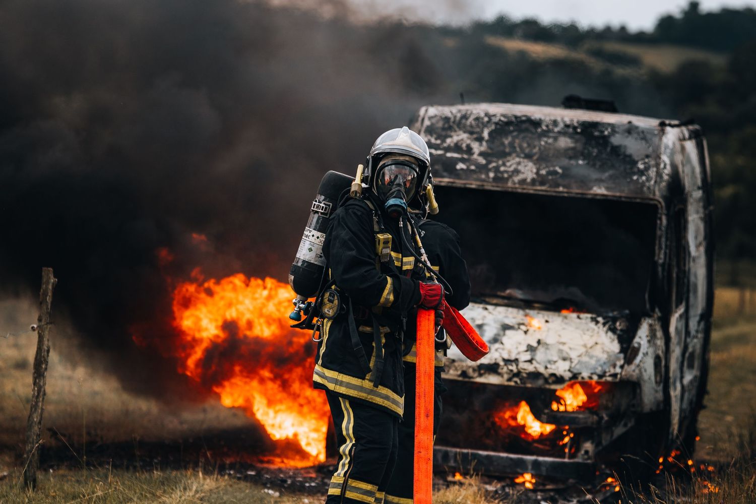Firefighter in full protective gear holding a hose battles a burning vehicle engulfed in flames outdoors.