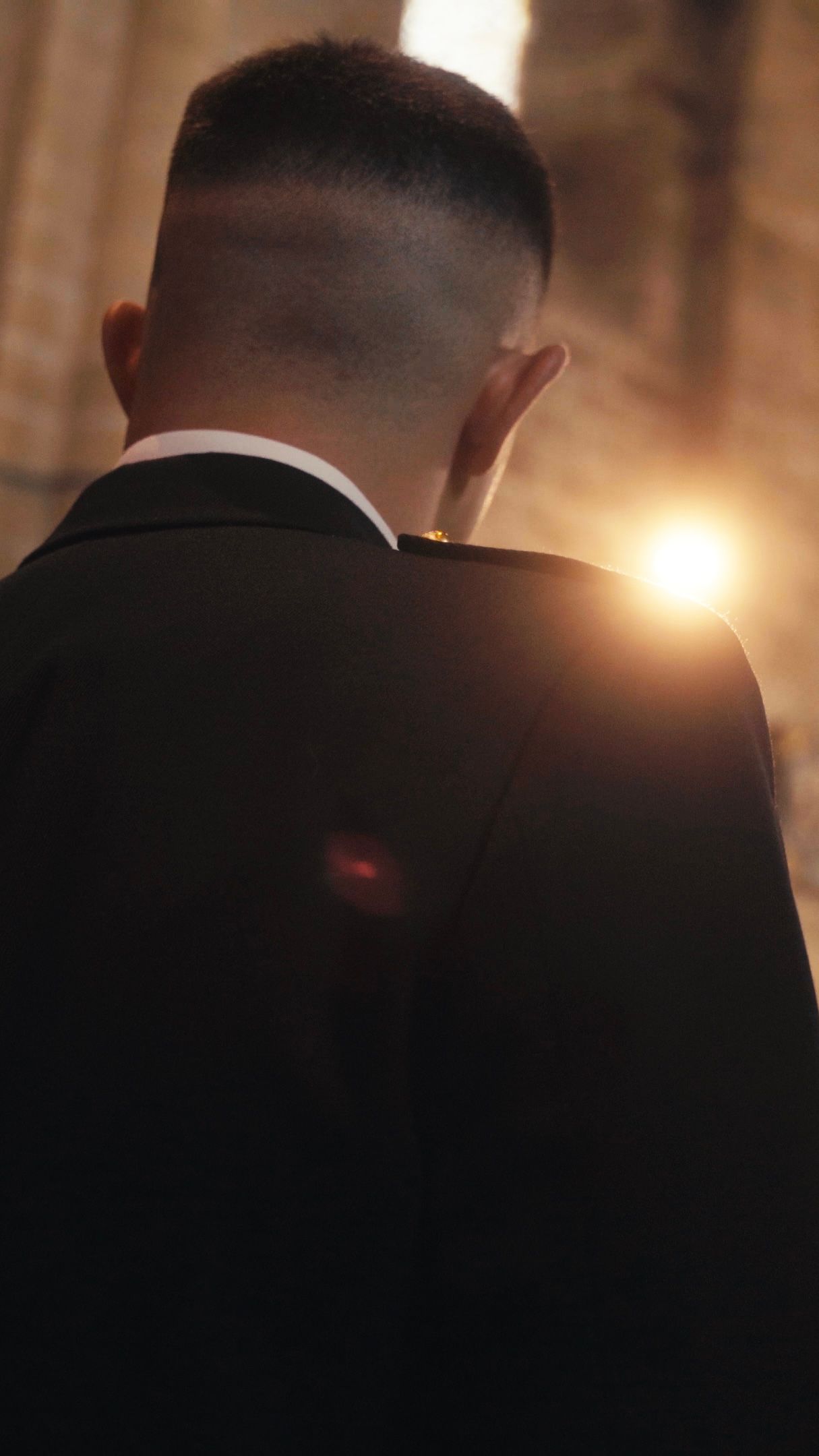 A man in a black suit stands with his back to the camera bathed in warm golden light indoors.