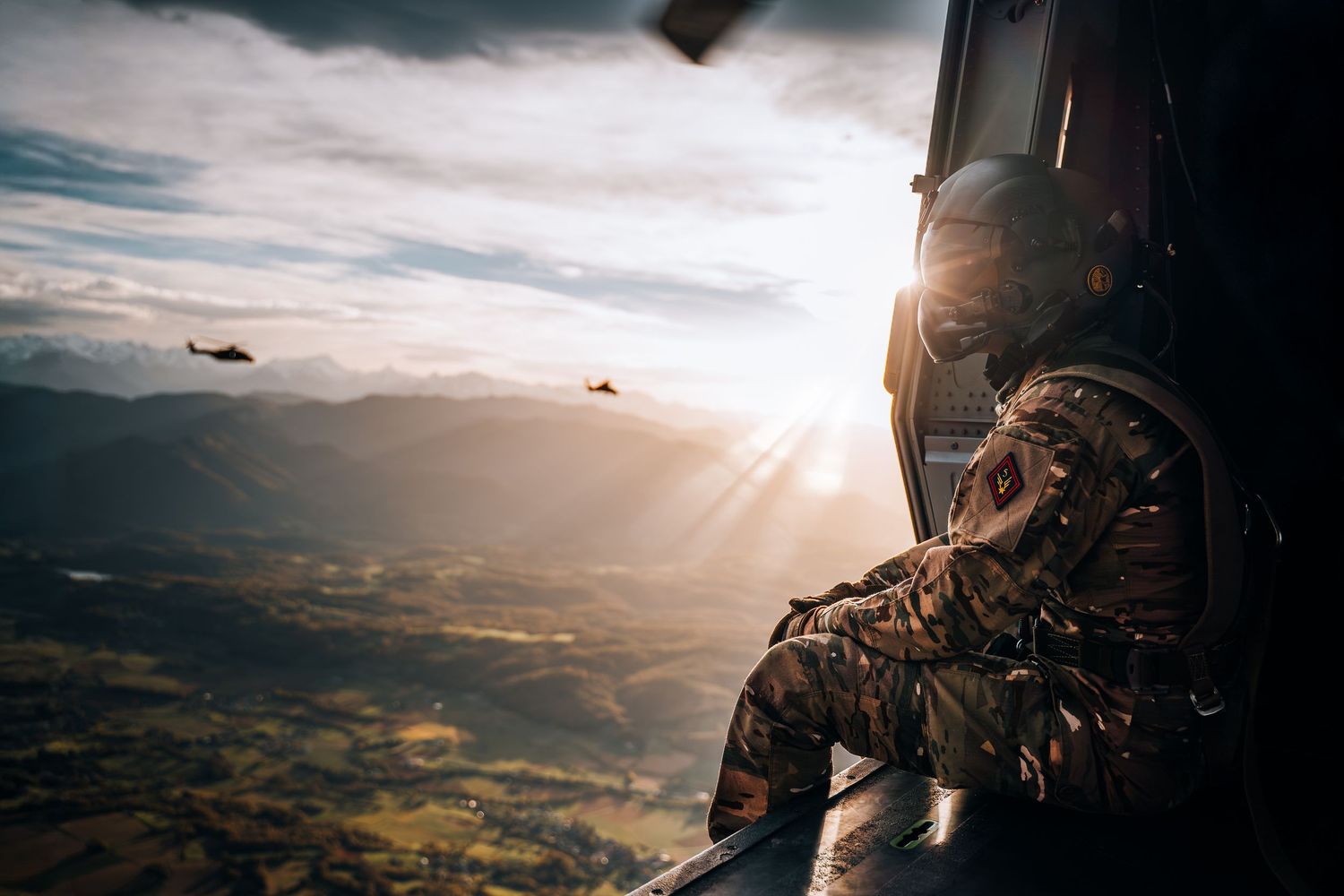 Military soldier seated in helicopter doorway watches other helicopters fly over sunlit valley.