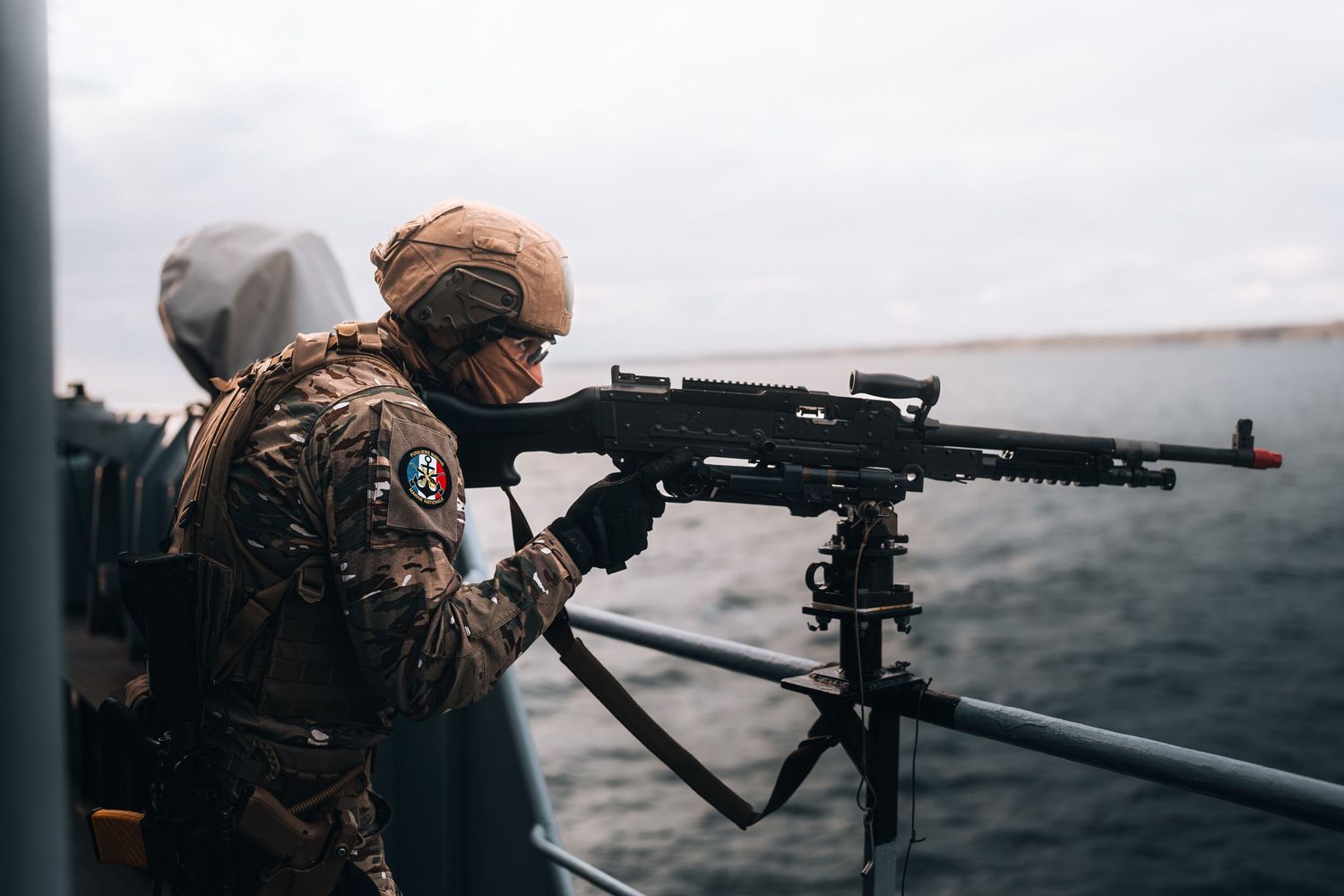 Military soldier in camouflage gear operates mounted machine gun on naval vessel at sea.